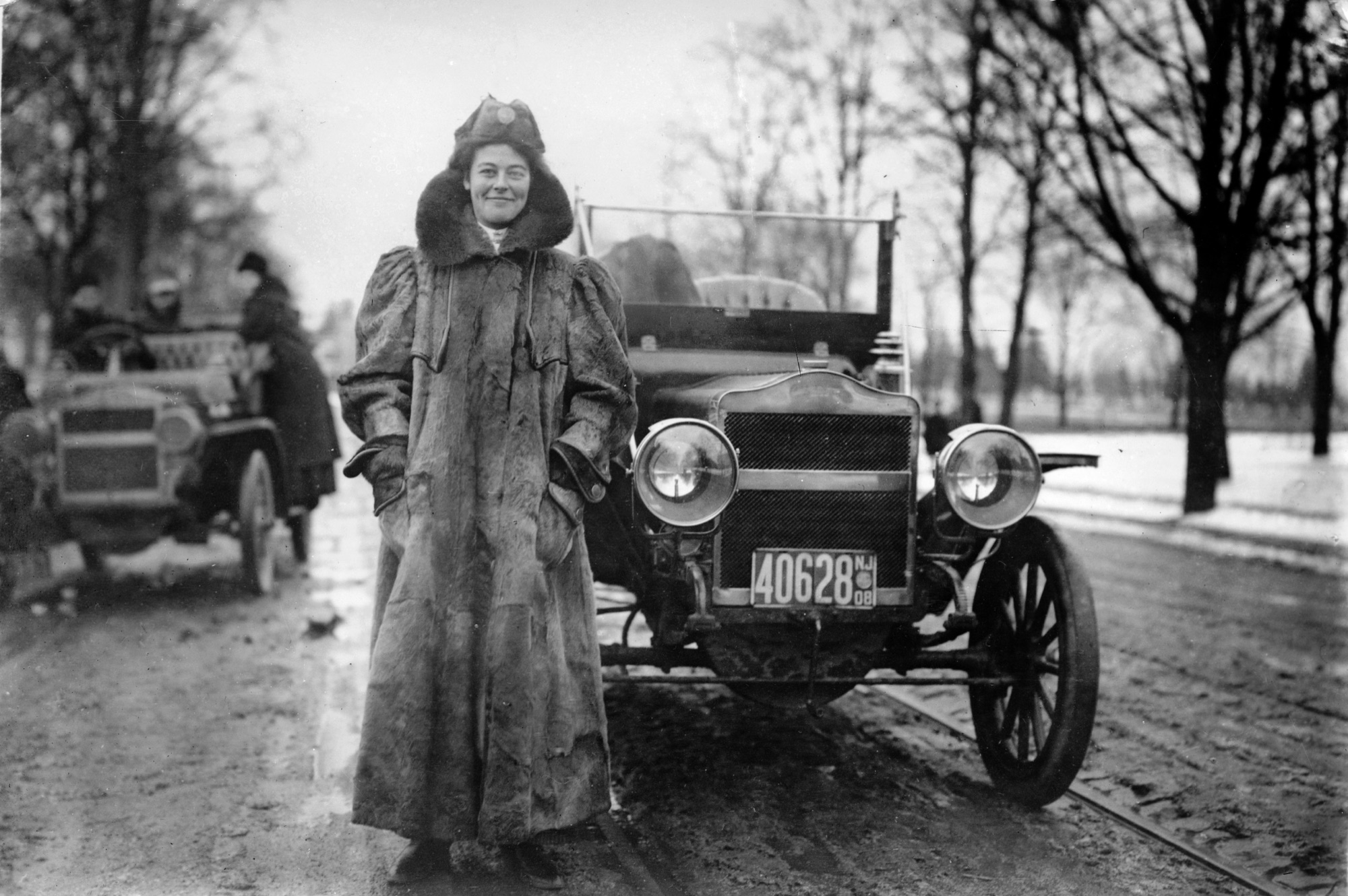A woman in a heavy fur-collared coat and fur hat stands beside an open-top touring automobile parked on a wet, tree-lined road in winter, a second early car visible behind her.