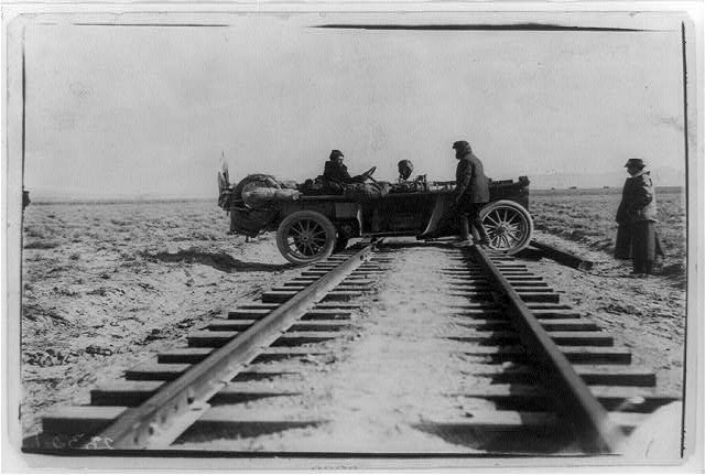 An open touring car from the 1908 New York to San Francisco automobile race, stopped astride a railroad track in open country with its crew standing beside it