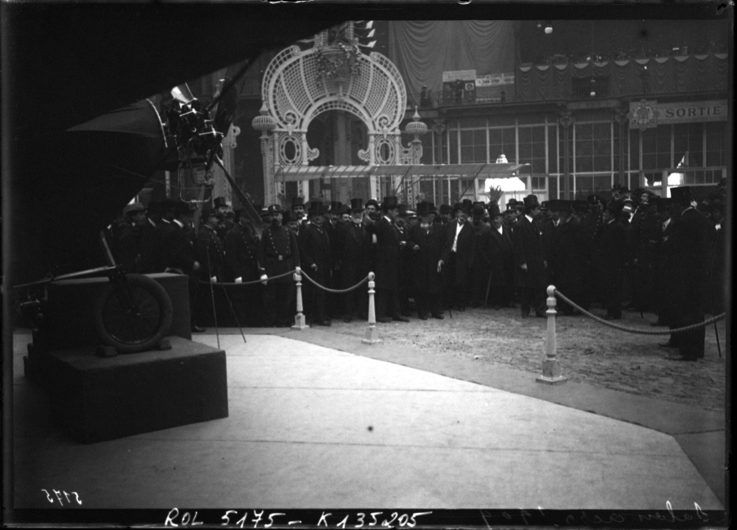 A 1909 photograph of top-hatted dignitaries at the Paris aeronautics salon, roped off around an early aeroplane displayed on a plinth, appraising a machine no one yet knew how to price