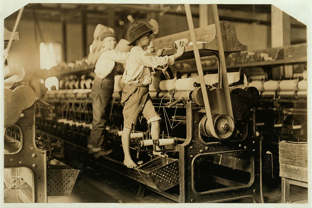 Two barefoot boys standing directly on the moving parts of a cotton spinning frame inside a mill, reaching up to tend rows of bobbins and thread, with rows of machinery stretching into the background.