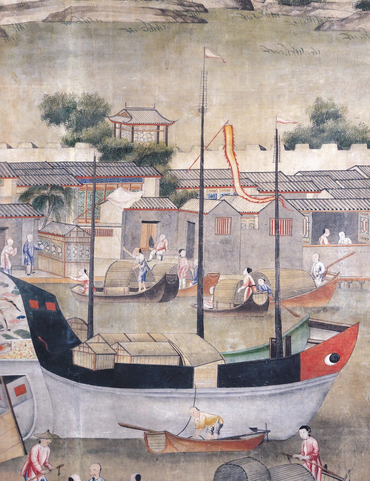 A large cargo junk with a black, red, and white painted hull and an eye motif on the bow, moored at a walled river town while smaller sampans load and unload cargo baskets alongside it.