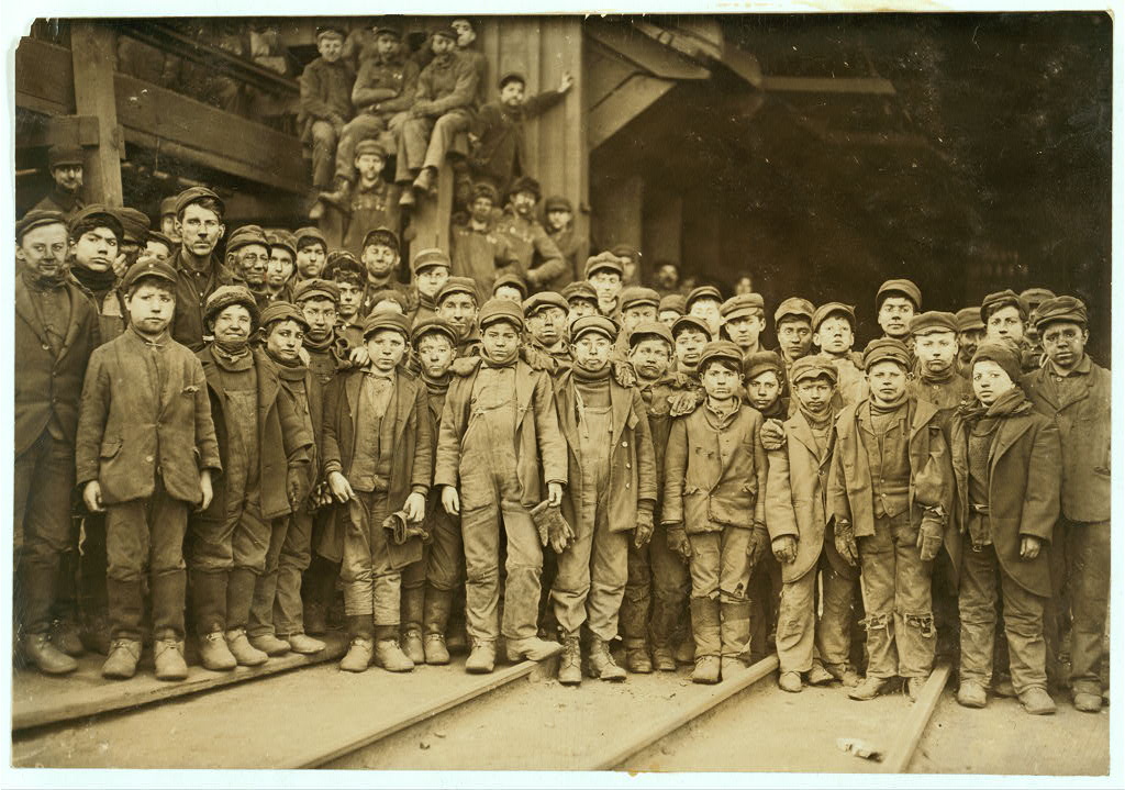 A large group of coal-dust-covered boys and young men, some standing and some seated on machinery steps behind them, posed outside a coal breaker building, wearing worn jackets and caps.
