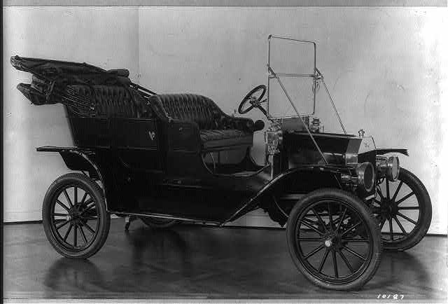A studio photograph of an early Ford motorcar in profile, an open touring car with a folded top, wooden spoke wheels, and a tiller-style steering wheel, against a plain backdrop.