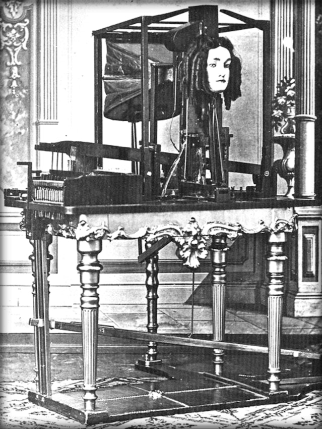 A photograph of Joseph Faber's Euphonia speaking machine of about 1846: a keyboard and bellows mechanism mounted on an ornate table, behind which a mask of a human face with long hair is fixed in a wooden frame, connected by rods and levers to the keys the operator pressed to make it form words.