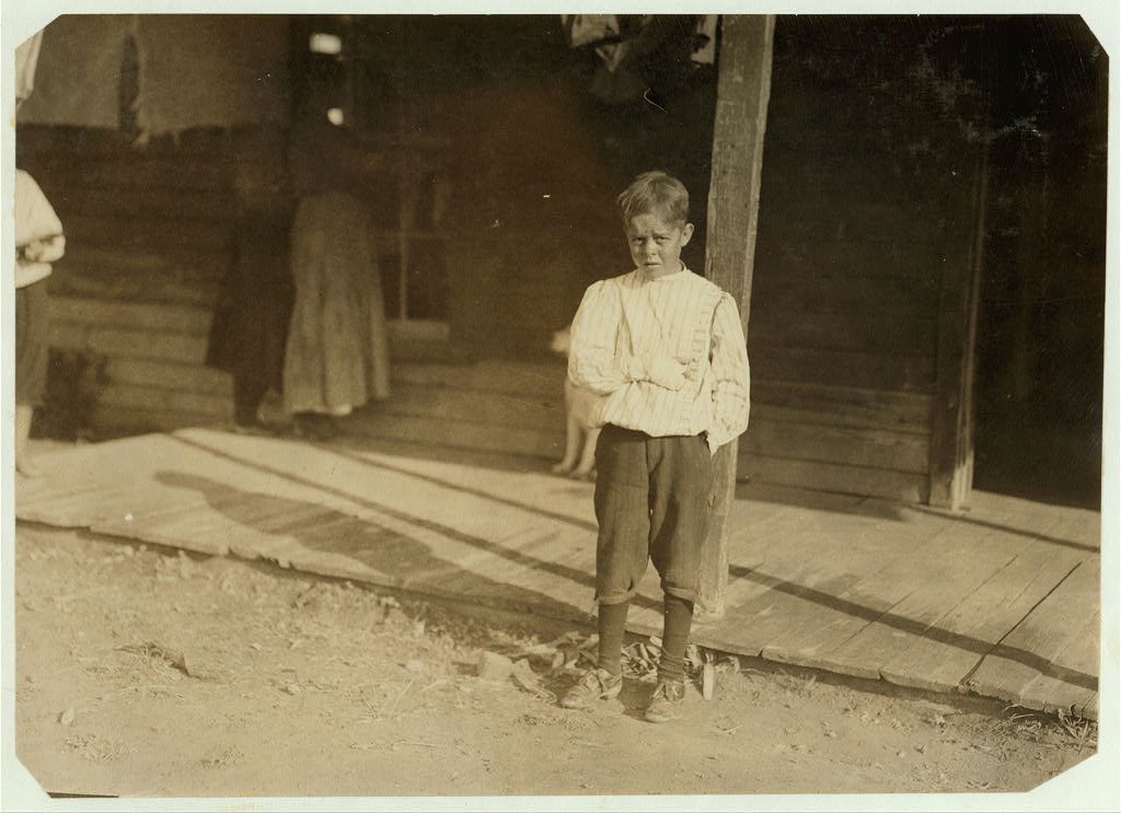 A young boy in a striped shirt and dark trousers stands outdoors on a wooden porch, his injured hand wrapped and held close against his body, two figures visible in the doorway behind him.