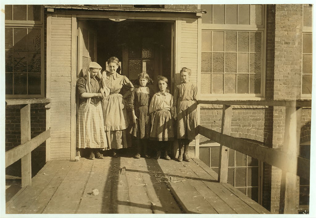 Five young female mill workers, dressed in period pinafores and aprons, standing together in a factory doorway on a wooden loading platform, with the brick mill building and multi-pane windows behind them.