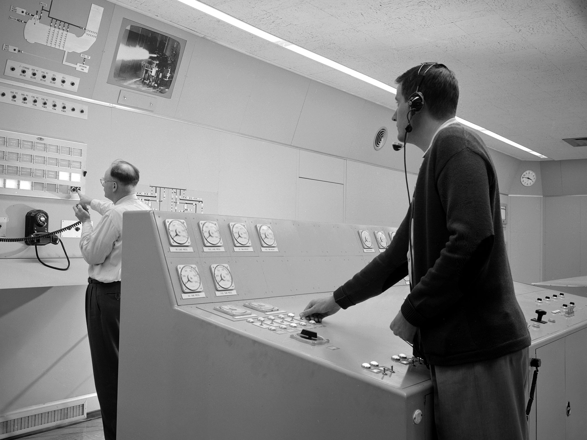 Two test engineers in a control room, one wearing a headset and operating a console with rows of round gauges and a keypad, the other standing at a wall panel of switches and indicator lights, a wall clock visible above.