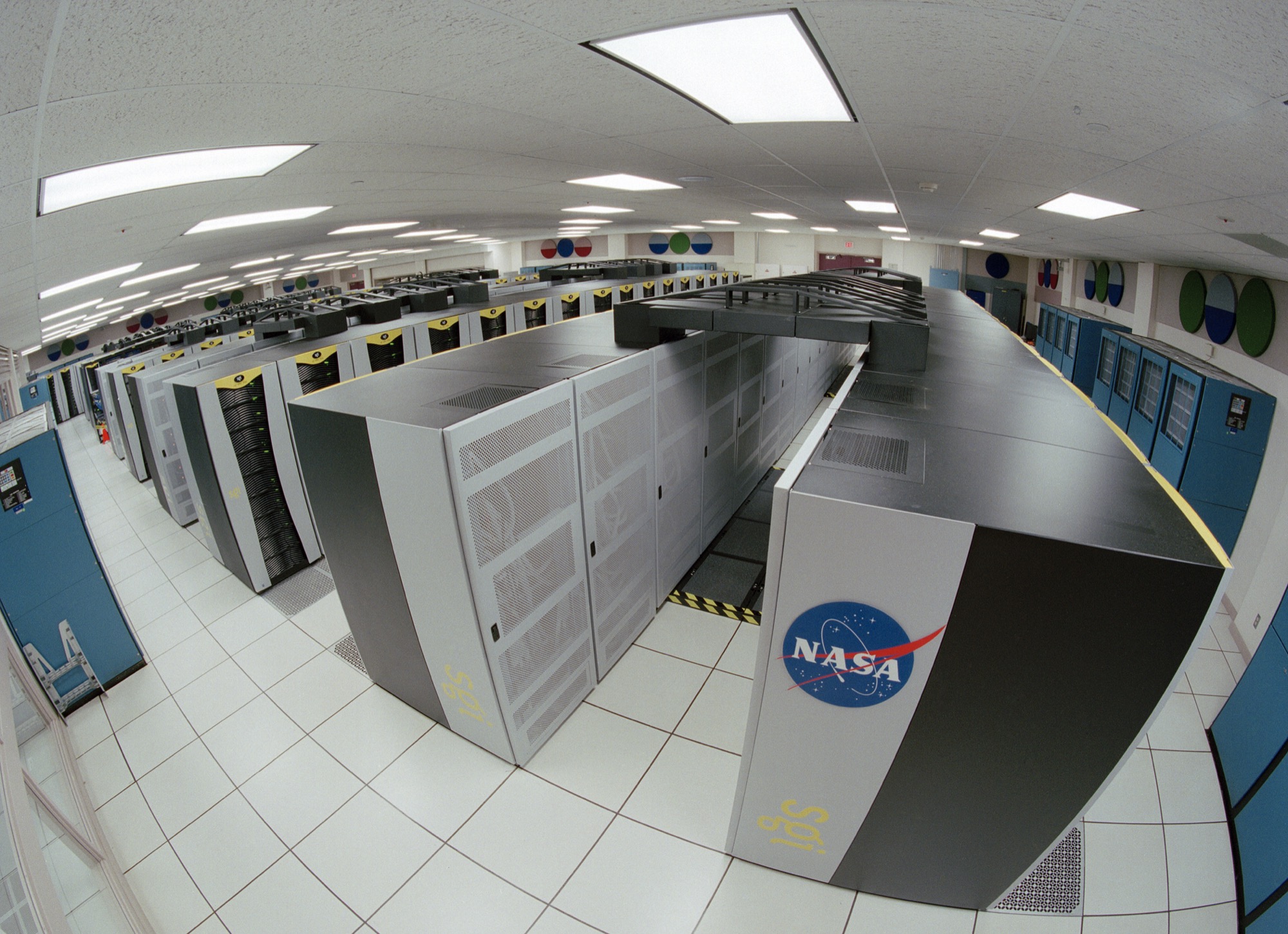 Wide angle view down an aisle of supercomputer cabinets in a NASA data center, with a NASA logo on the nearest unit.