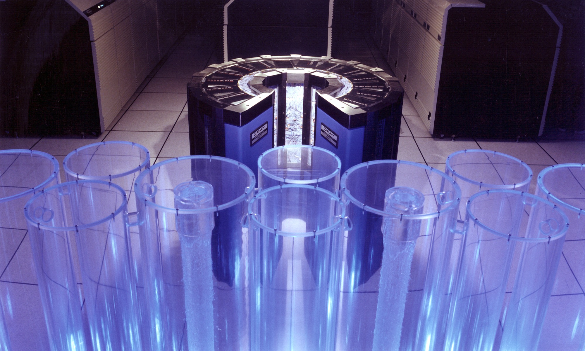 Cray-2 supercomputer in its distinctive curved cabinet, lit blue, with clear cooling columns in the foreground of the machine room.