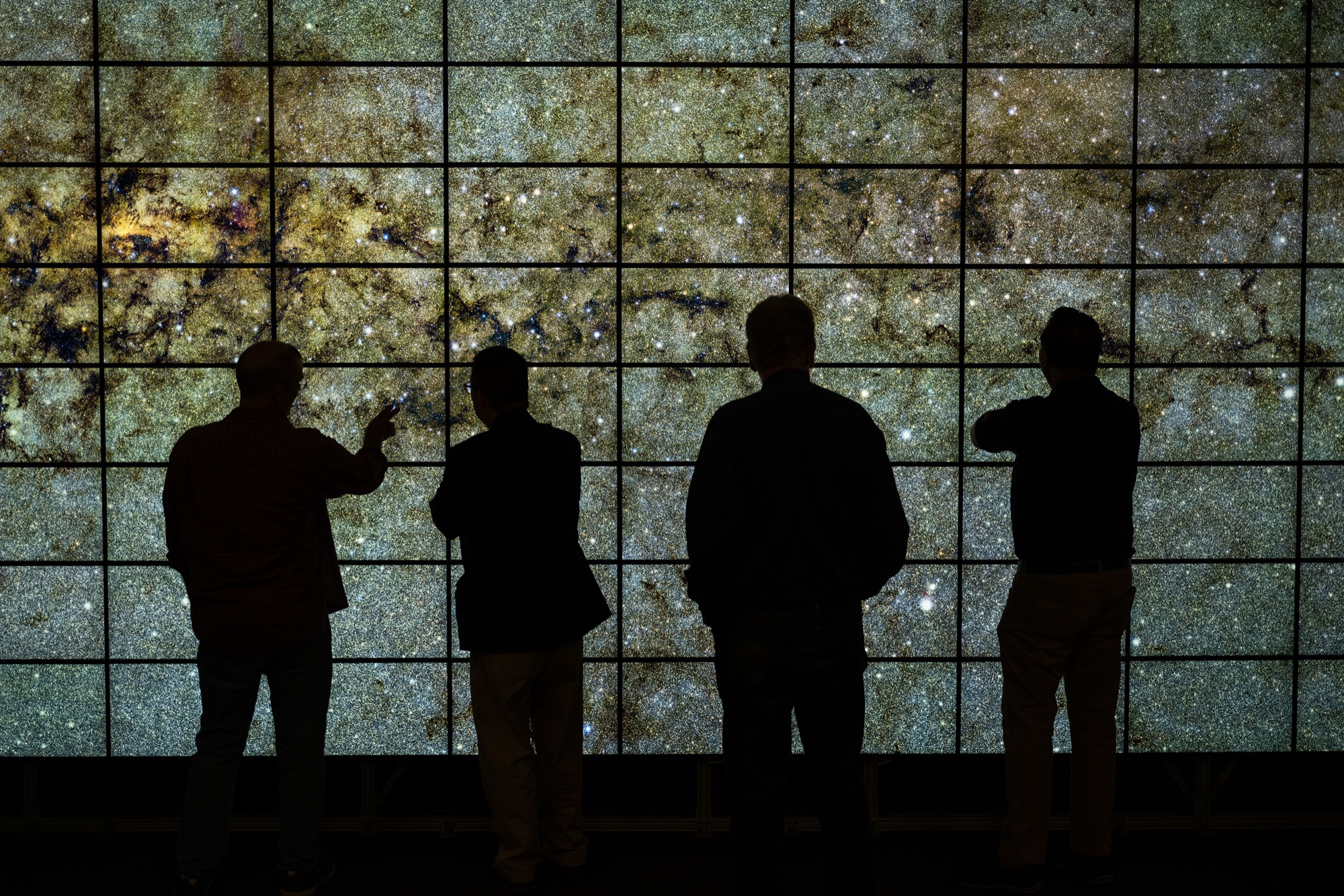 Four people stand in silhouette before a large wall of screens tiled into a grid, displaying a dense field of stars and data overlays, in a darkened visualization room.