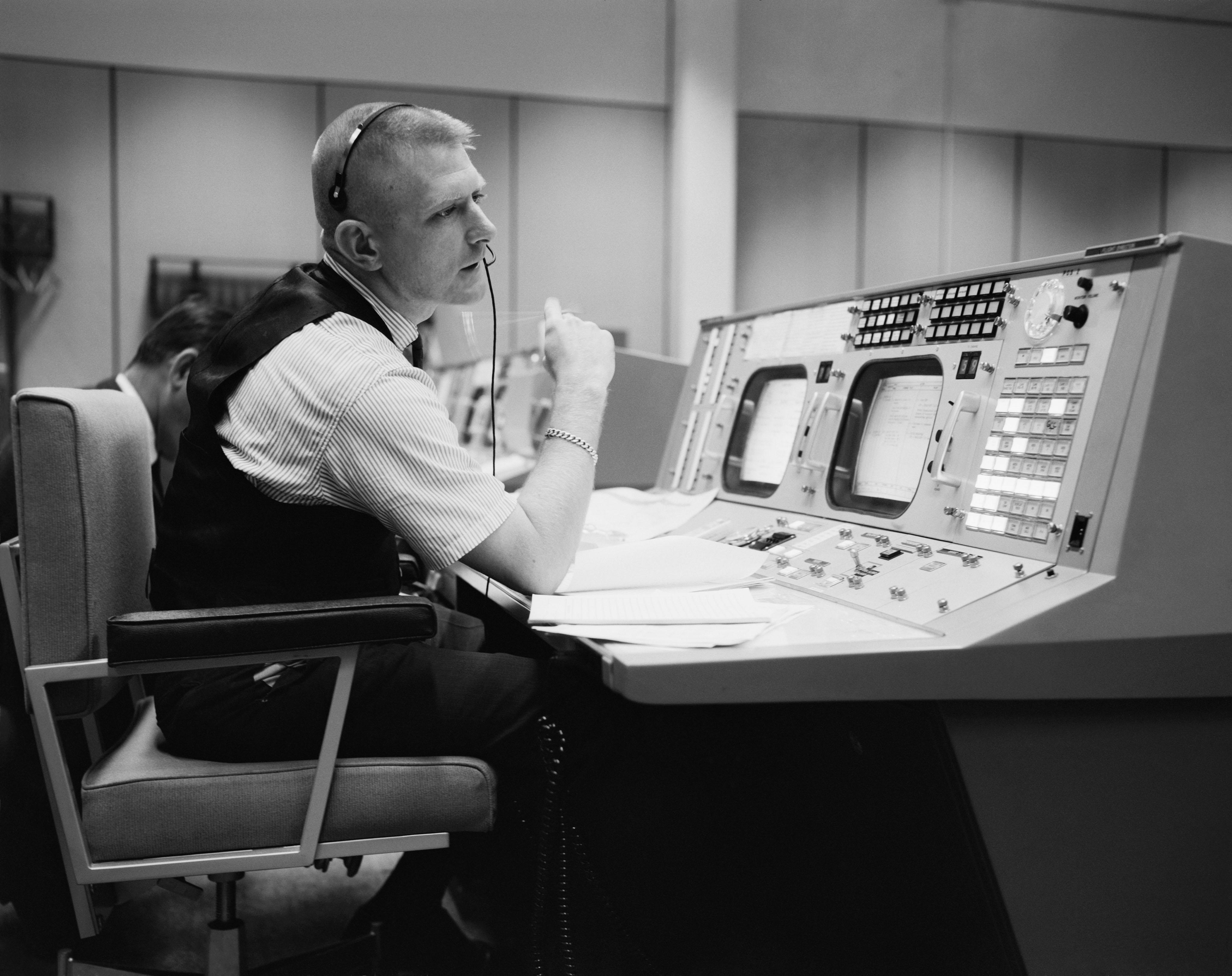 A NASA flight director at a Mission Control console in 1965, headset on, watching banks of monitors, one hand ready to halt the mission the instant something goes wrong