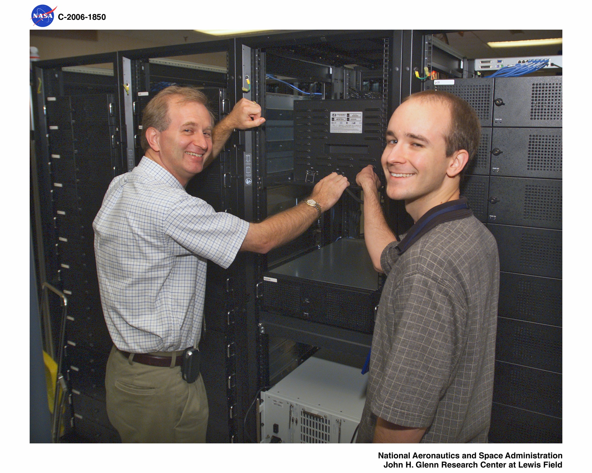 Two NASA engineers, one older man in a checked shirt and one younger man in a gray polo shirt, standing at an open equipment rack in a server room, both smiling toward the camera with hands raised near the rack door.