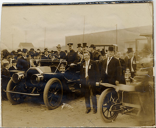A large open touring automobile parked on a dirt lot, surrounded by a dense crowd of men in top hats and bowler hats, with a second automobile visible in the foreground.